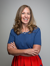 Photo of Janikke Klem from waist up, Caucasian female, long curled dark blonde hair, wearing blue short sleeve blouse tucked into red skirt and ring linked necklace