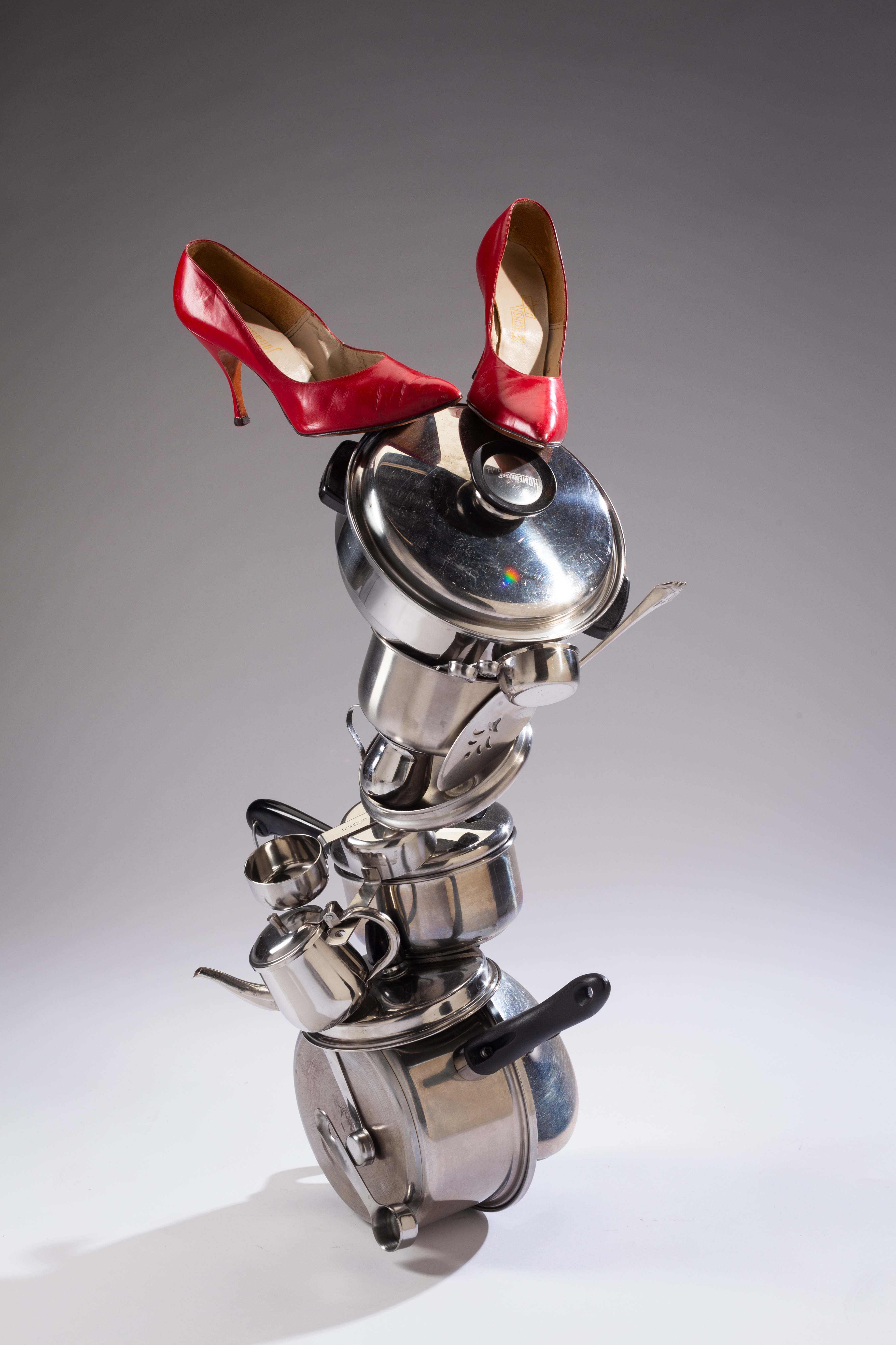 Photograph of a sculpture featuring a stack of pots and pans with a pair of red high heels resting on top.