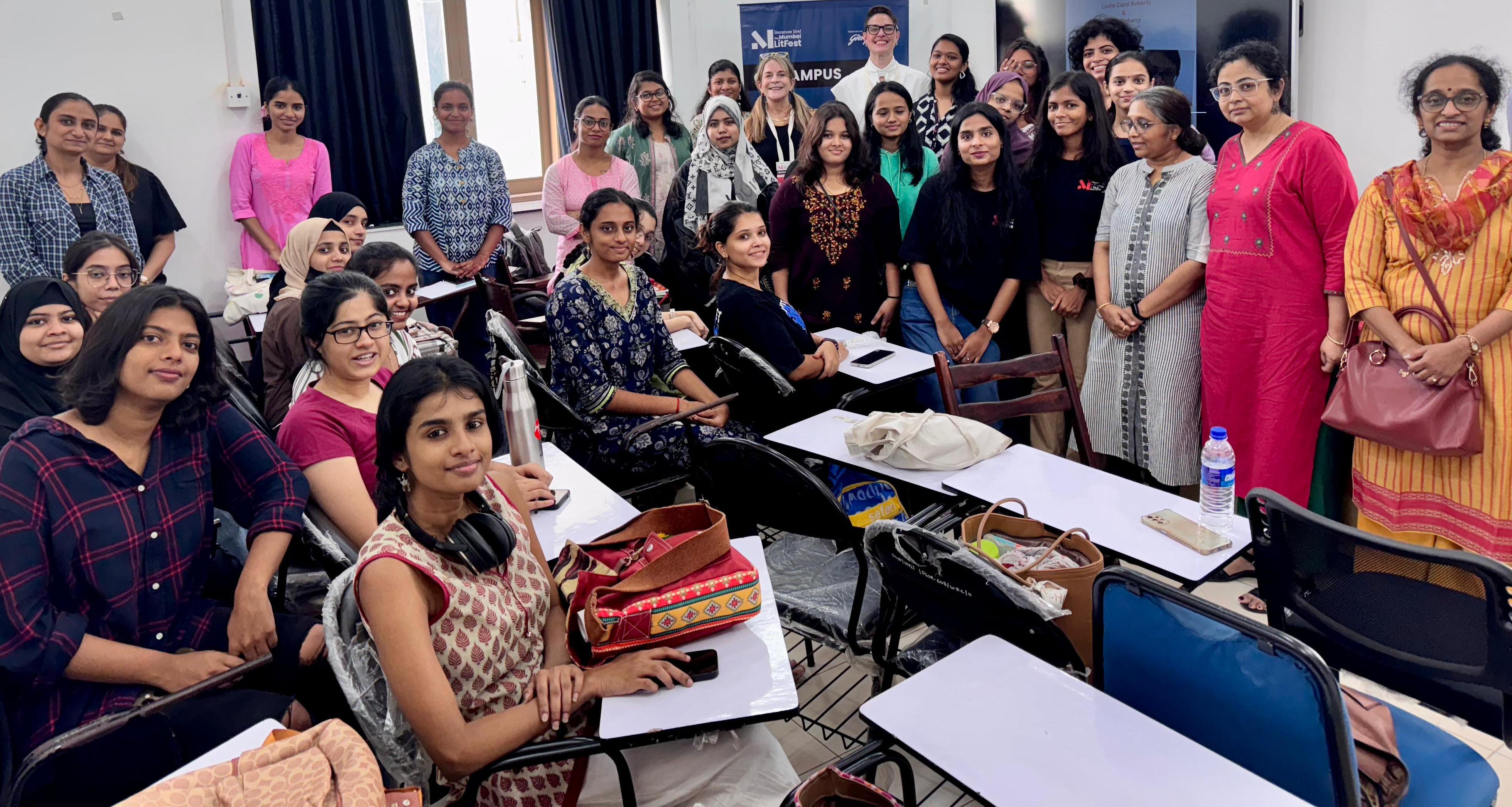  A diverse group of women, along with a few instructors, pose for a group photo in a classroom setting. Some are seated at desks with notebooks, bags, and water bottles, while others stand behind them, smiling at the camera. The room has white walls, black curtains, and a banner in the background indicating an academic or workshop setting. The participants are dressed in a mix of traditional and modern clothing, reflecting a vibrant and engaged learning environment.