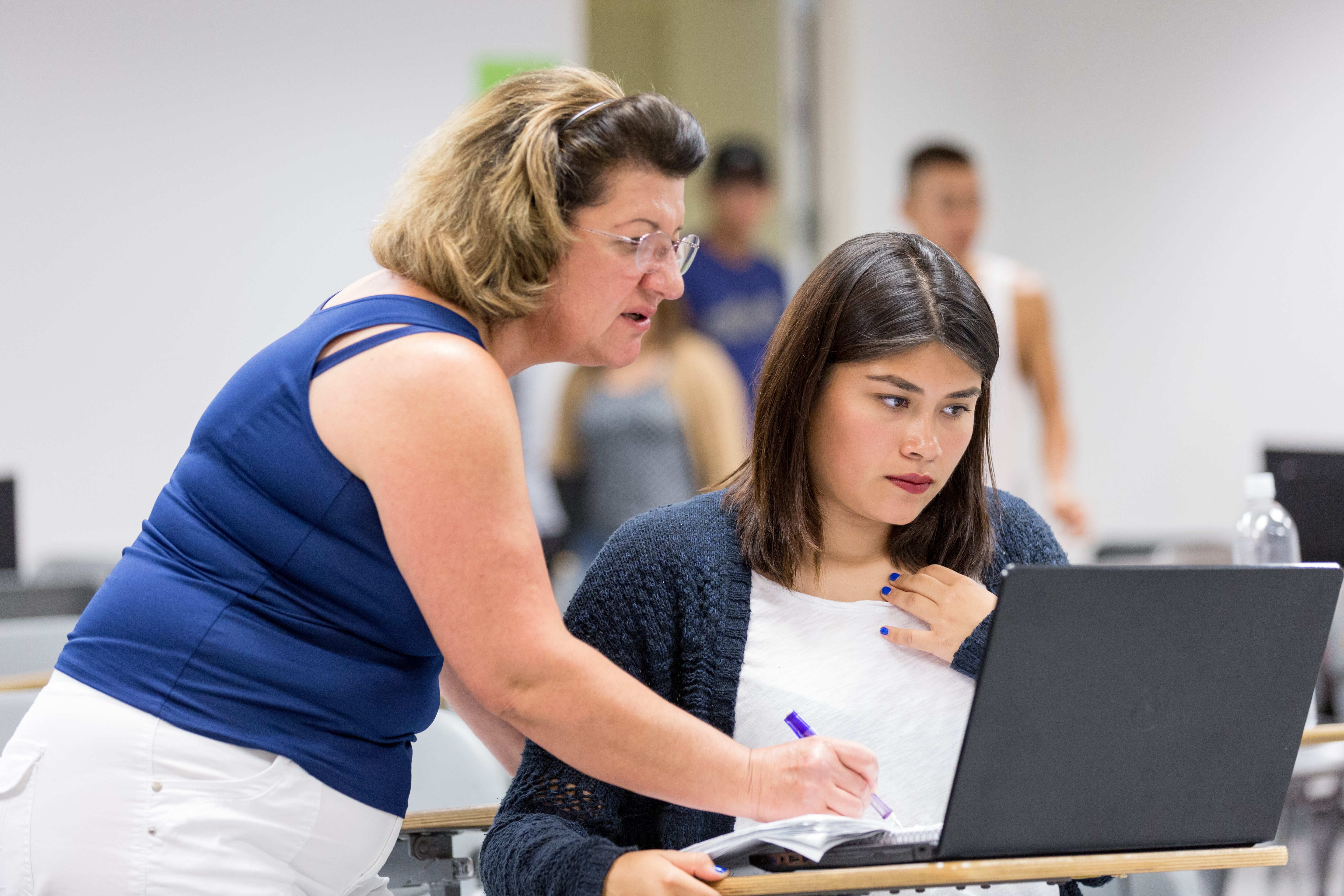 A teacher leans over to help a student with work on her computer and worksheet.