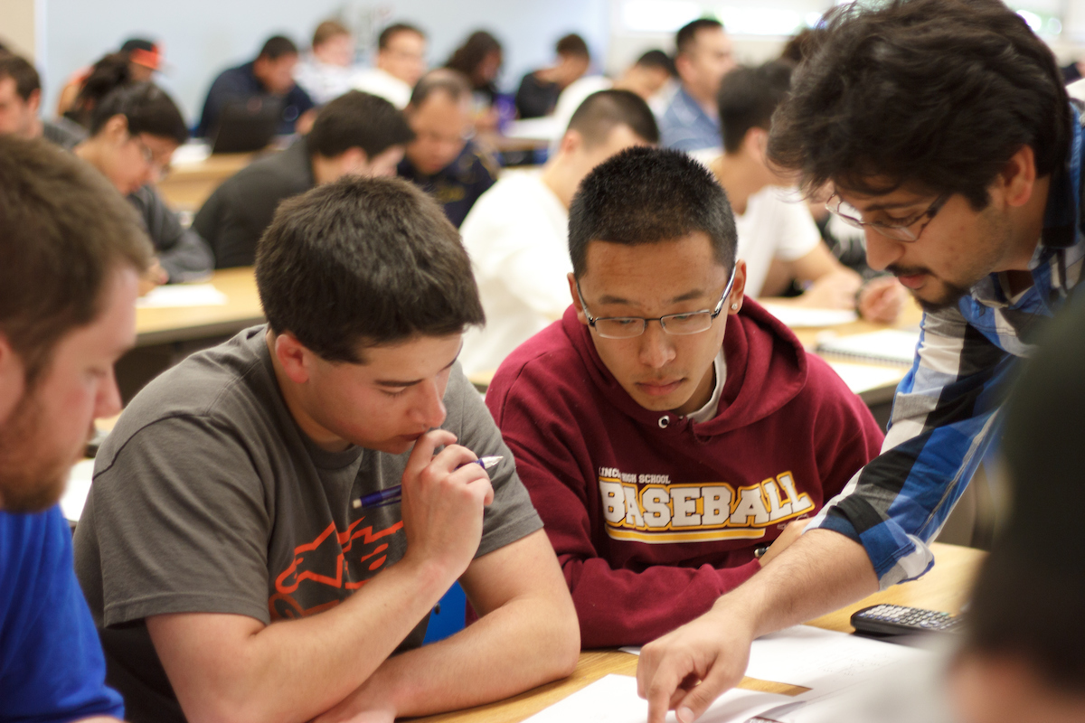 Four students sit inside a full classroom as they look at a paper on their desk.