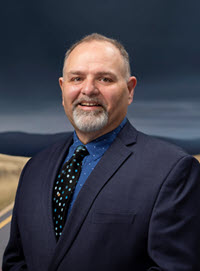 Professional portrait of David Renfro. He is an middled-aged gentleman with greying hair and an open smile, posed in a dark blue suit.