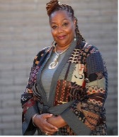 Black woman with dark brown hair, wearing an orange and brown, patterned suit jacket, smiling in front of a gray brick wall. 