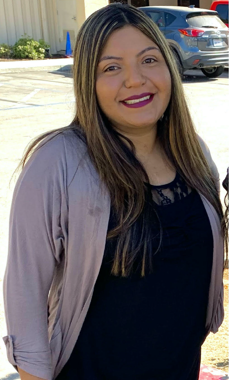 Hispanic woman with long, brown and blonde hair, wearing a gray cardigan and black shirt, smiling in a parking lot.