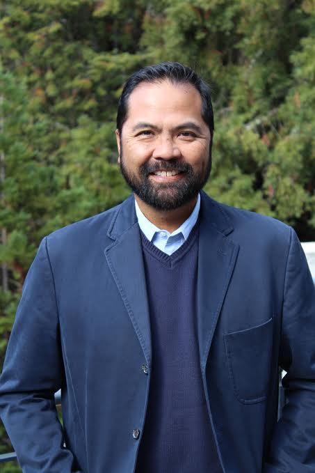 headshot of glenn gumin in blue blazer with greenery in background 